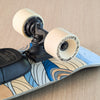 Close-up of a longboard skateboard with cream-colored wheels, black trucks, and a wood-grain deck with blue and beige wave patterns, resting on a light wood surface.